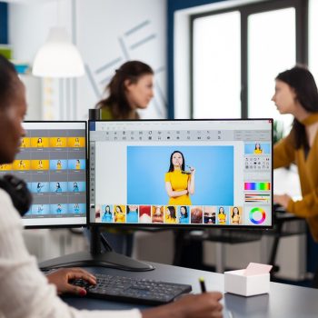 Black retoucher woman analysing images on pc with two monitors and retouching digital assets with stylus pencil. African project designer using photo editing software sitting in creative agency office